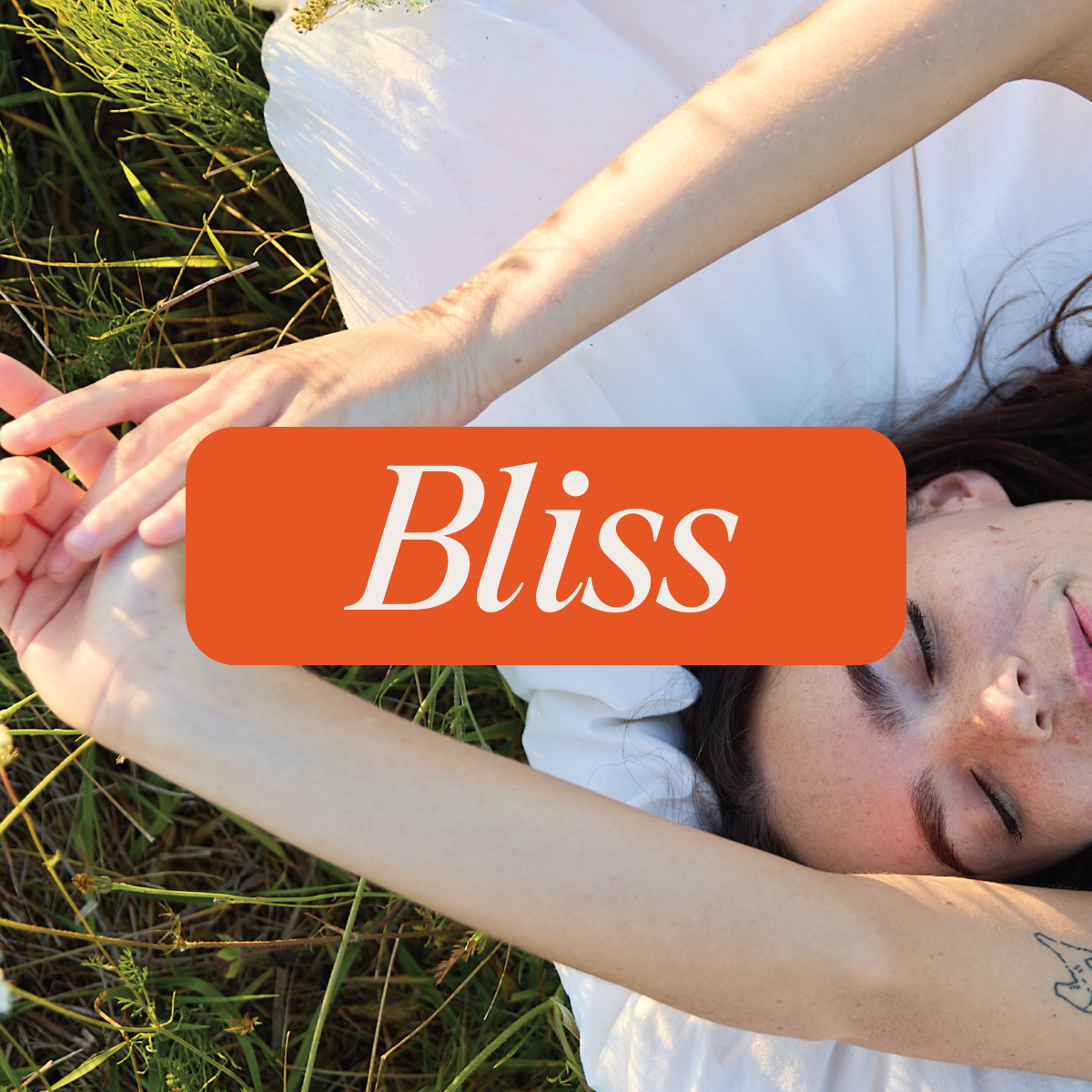 Person lying on grass with eyes closed and arms stretched overhead, dressed in white, with the word 'Bliss' displayed on an orange banner—suggesting a serene, peaceful moment.