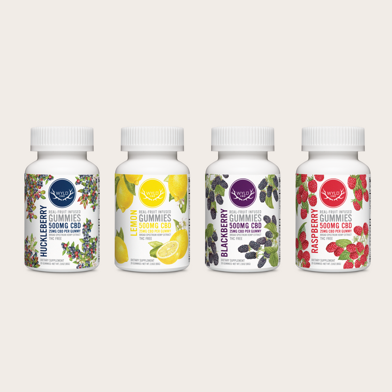 Four white bottles of Wyld Gummies 500mg CBD in different flavors (Huckleberry, Lemon, Blackberry, and Raspberry) lined up on a limestone background.