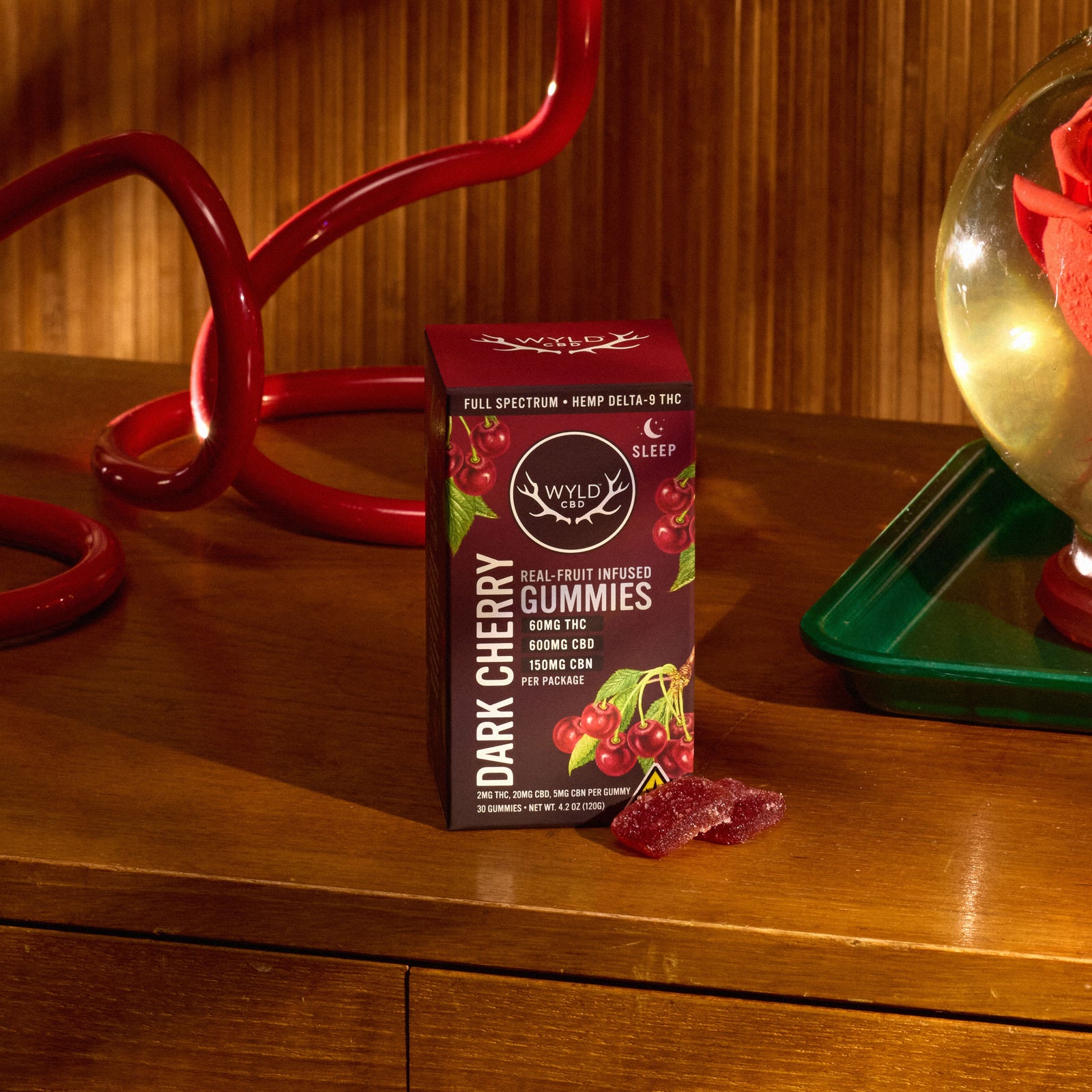 A box of Wyld Dark Cherry Sleep Gummies sits on a wooden table next to a red decorative object, with a few loose gummies in front of the package.